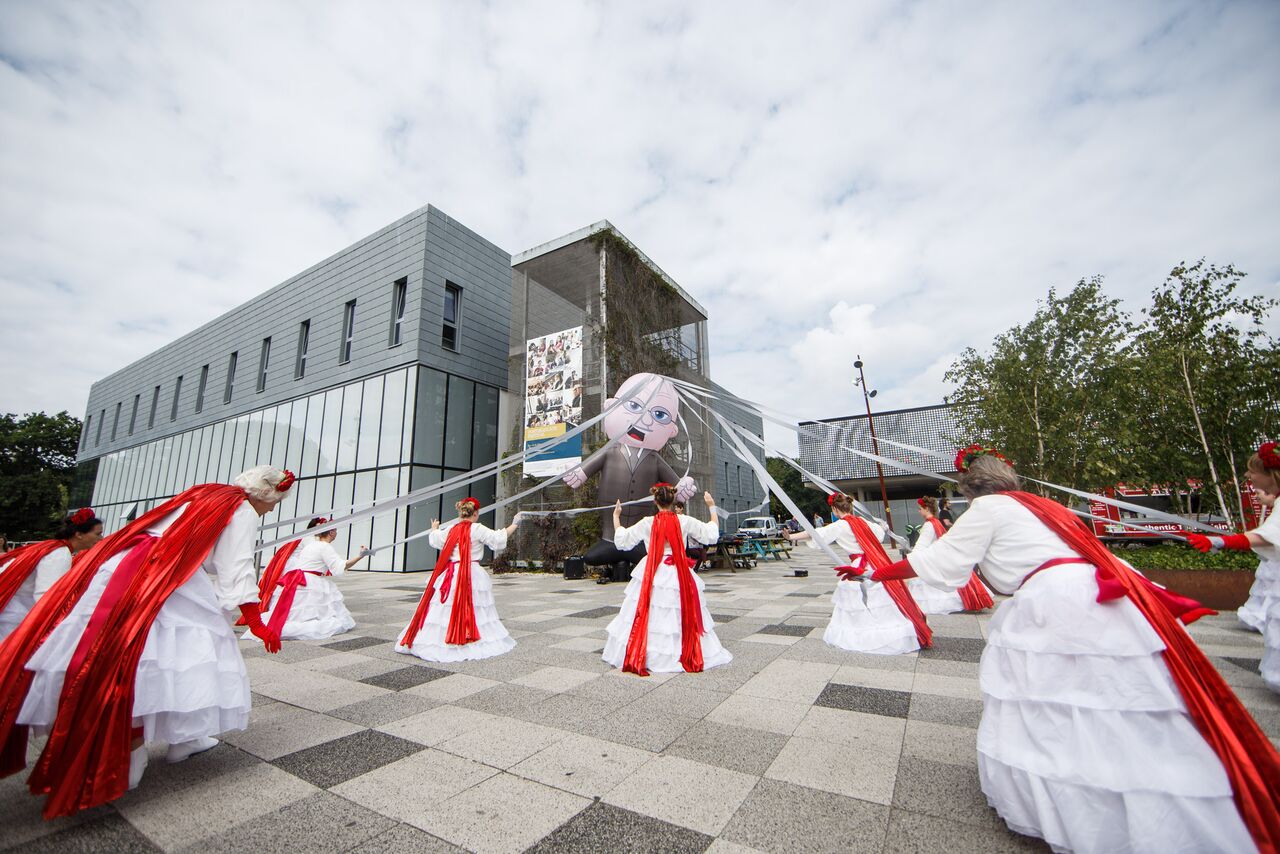 A group of dancers wearing white dresses with long red capes perform a routine outside a modern building, holding white ribbons that stretch outward toward a large, caricature-like inflatable puppet of a man positioned in the background.