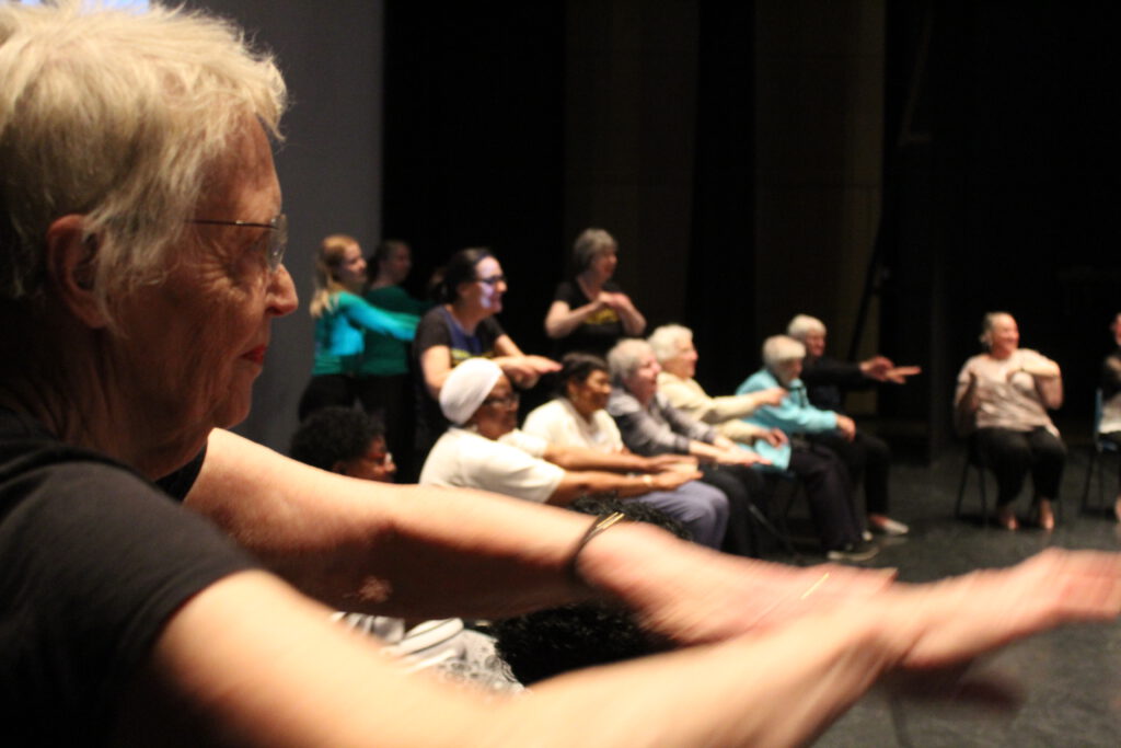 A group of older people sit and stand with their arms outstretched.