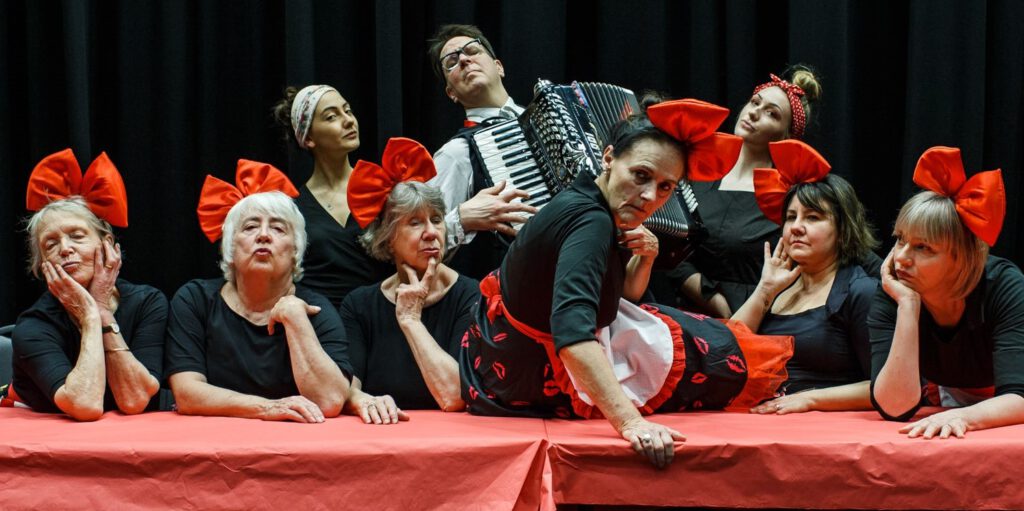 6 women with big red bows in their hair, flowery skirts and white aprons sit at a table and strike a pose while 3 musicians, including 1 man playing an accordion strike a pose in the background.