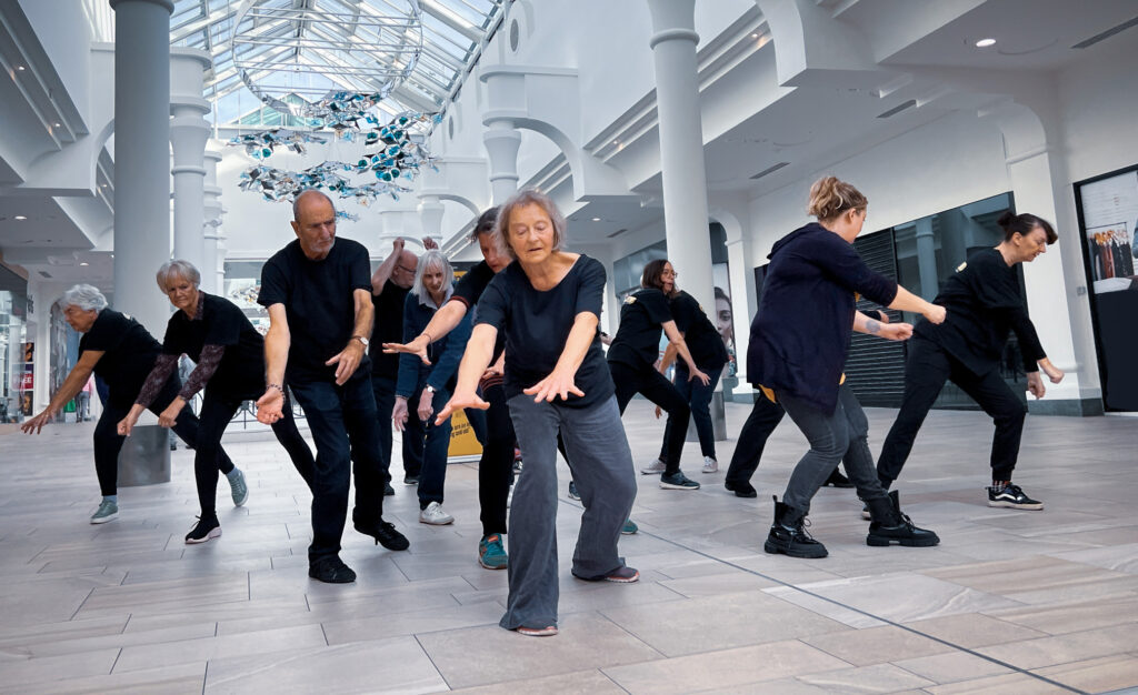 A group of men and women with arms outstretched and dressed in black dance in a shopping centre.