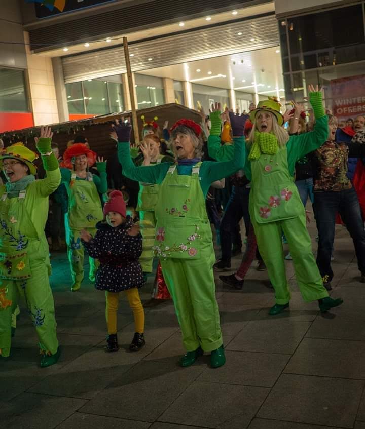 a group of women dressed in green dungarees raise their arms in surprise. A child with a red bobble hat stands next to them and does the same.