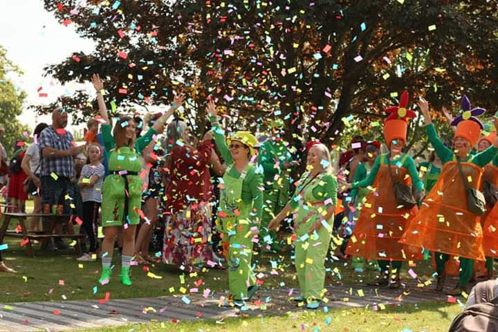 women in green dungarees and some dressed as flowerpots stand outside as confetti falls around them.