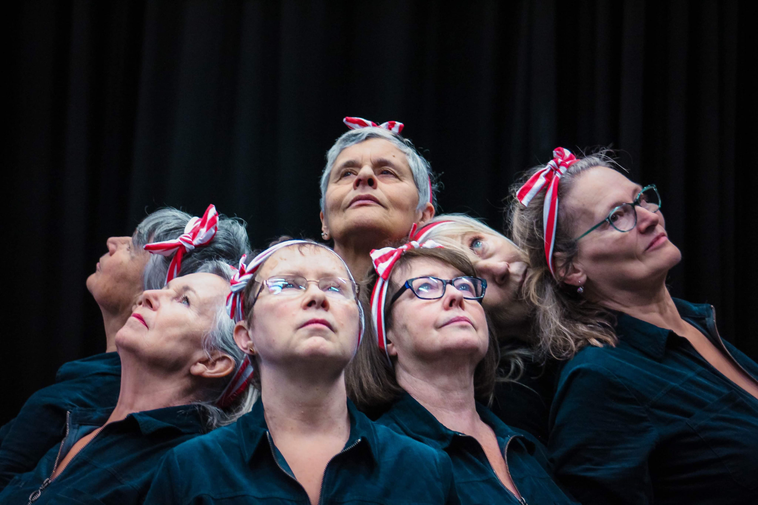 Six female performers in dark outfits and red-and-white headbands pose tightly clustered together, looking upwards during a performance.