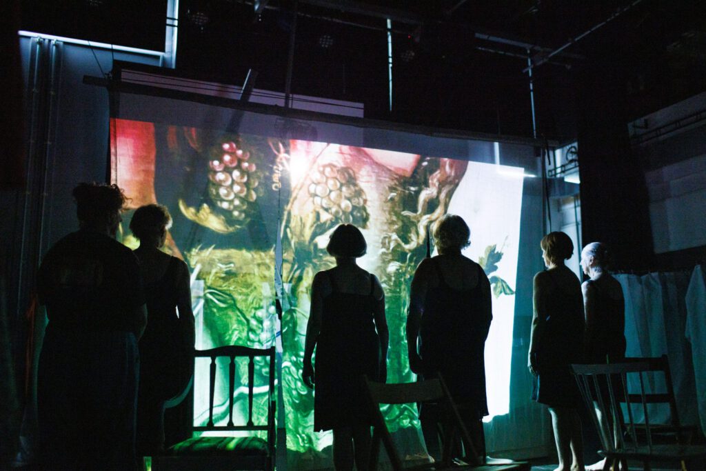5 women stand silhouetted with their backs to us in front of a large projected image.