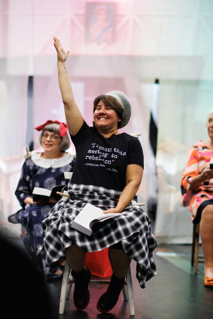 A woman in a checked skirt, sits on a stool and smiles while putting her hand up.