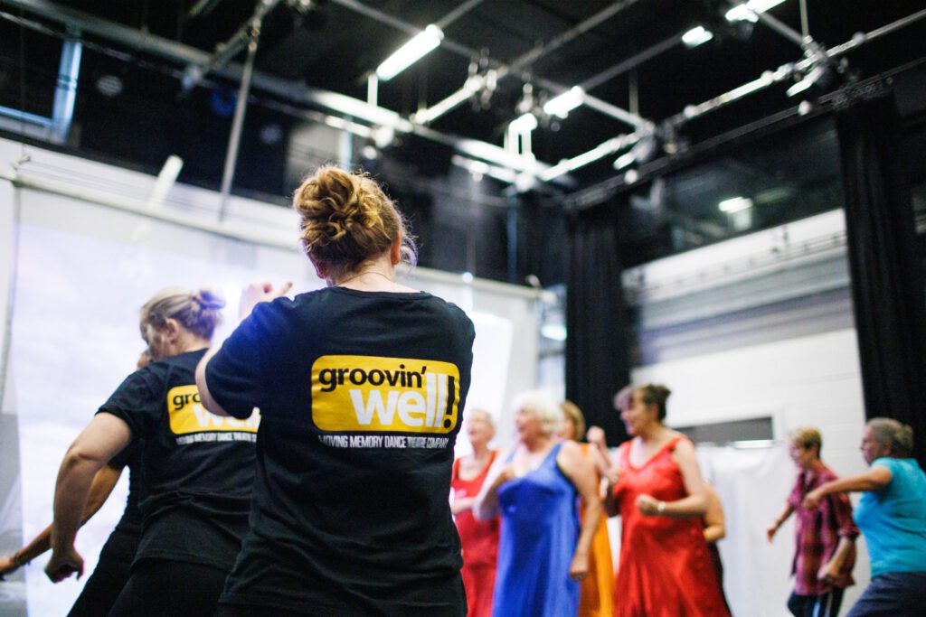 Two women with their backs to us, wearing Grooving Well T-shirts dance with a group of people in coloured clothing.