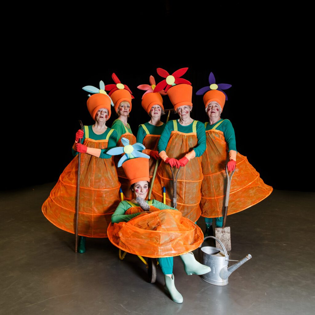6 women dressed as orange flower pots with flowers on their heads pose for a photo. They hold spades and one sits in a wheelbarrow next to a watering can.