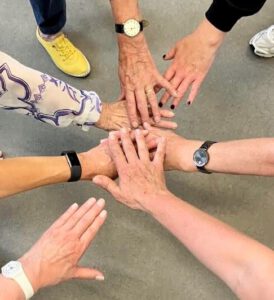 Moving Memory team members and participants reach inwards to place their hands together in a huddle against a grey floor.