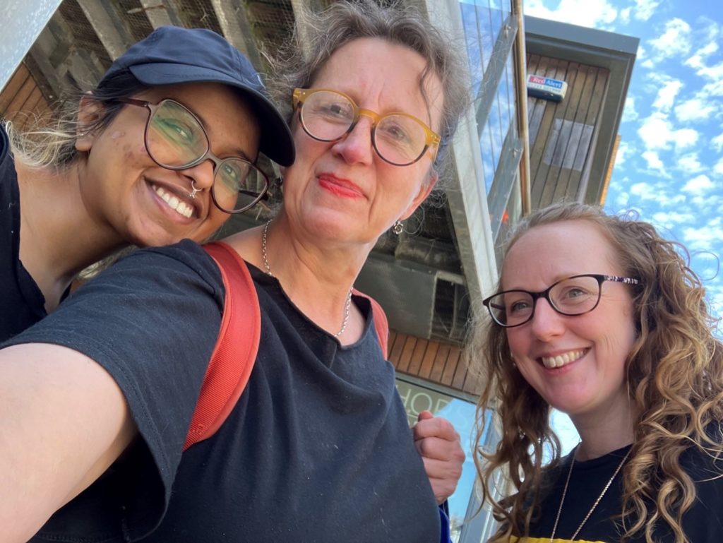 Three Moving Memory team members take a close, smiling selfie together outside under a bright sky.
