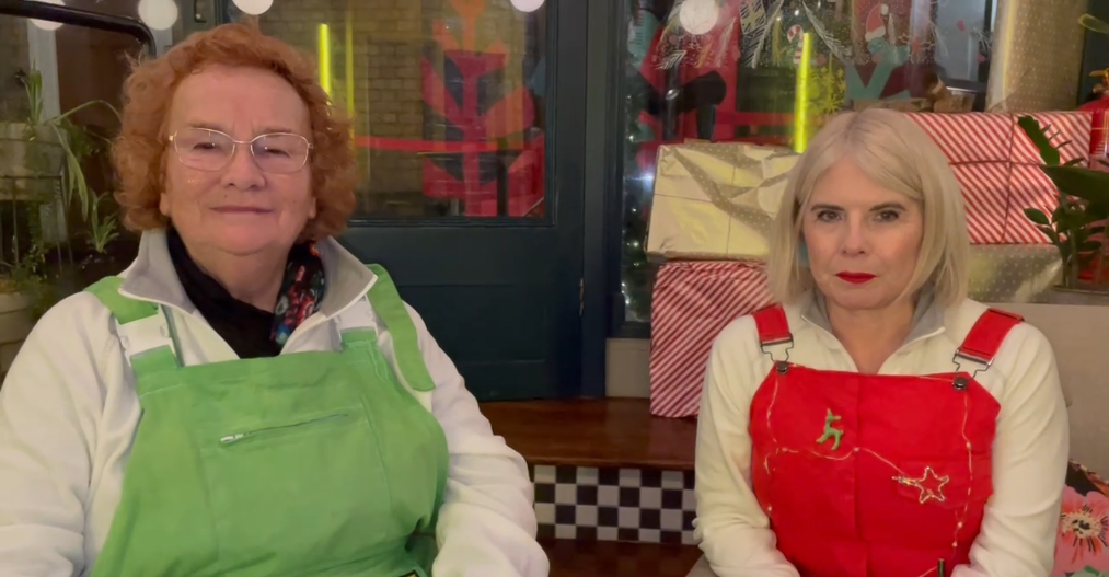Two women sit side by side in festive dunagrees—green on the left and red on the right—smiling in front of a window display filled with wrapped Christmas presents.