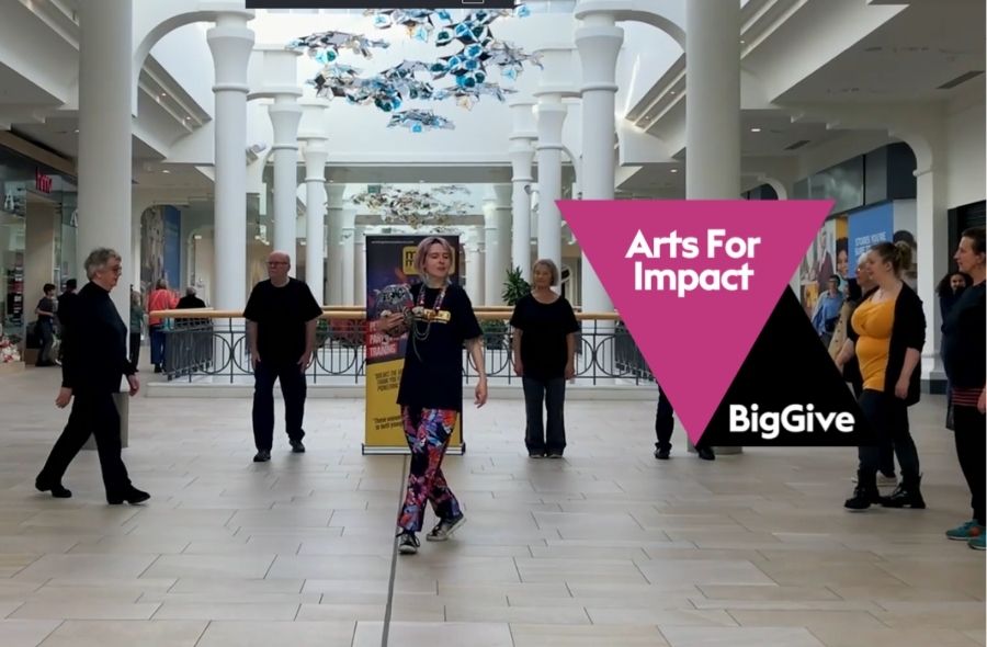 A group performs a dance piece in a bright shopping centre concourse led by a instructor in colourful patterned trousers. Overlaid graphics read "Arts For Impact" on a pink triangle and "BigGive" on a black triangle.