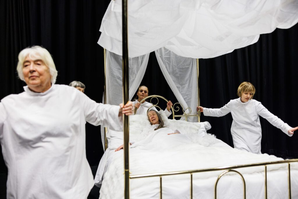 4 women dressed in white, push a brass 4 poster bed while a woman lies in the bed fast asleep under white sheets.
