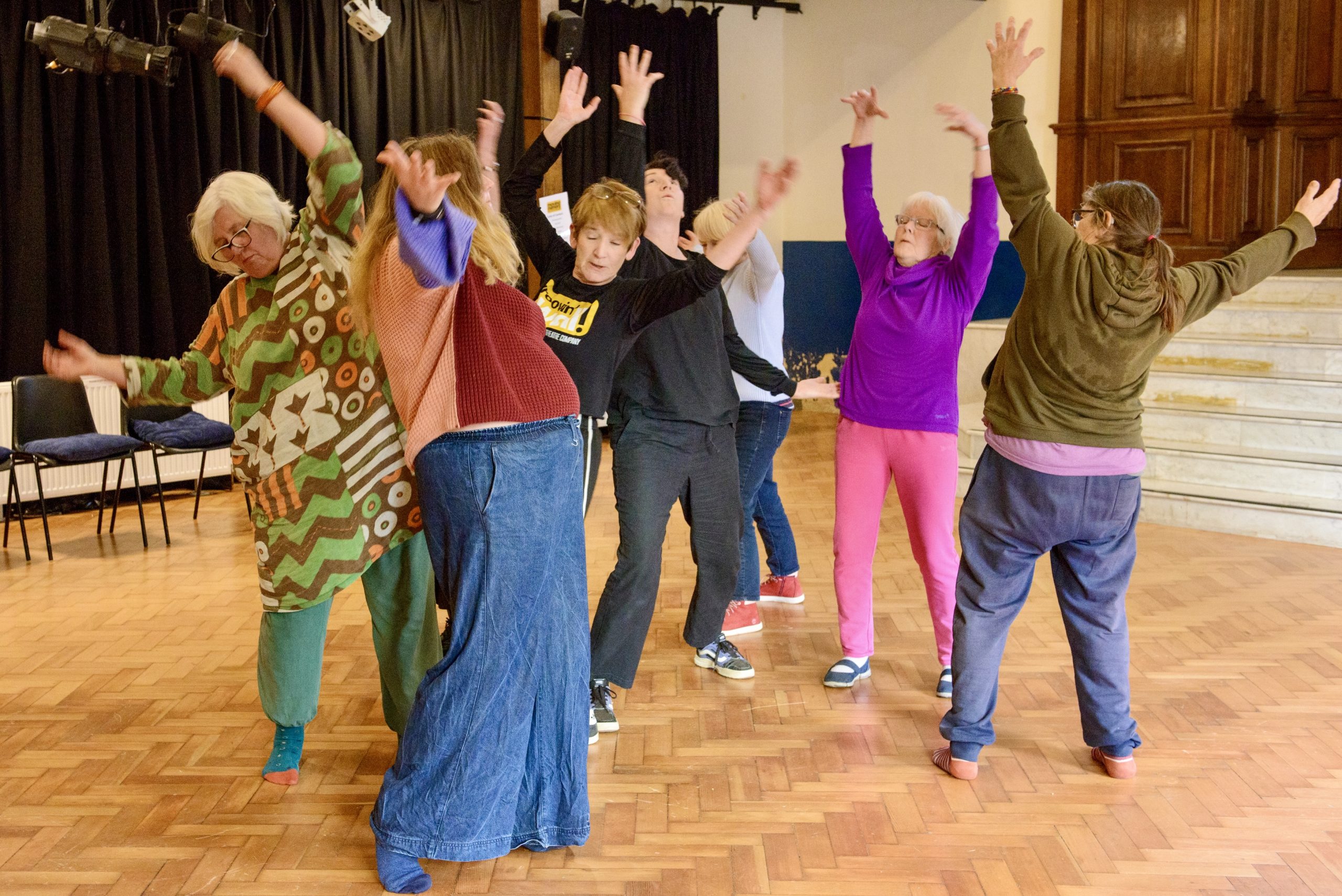 A group of people dressed in colour clothing dance in a hall with arms outstretched.