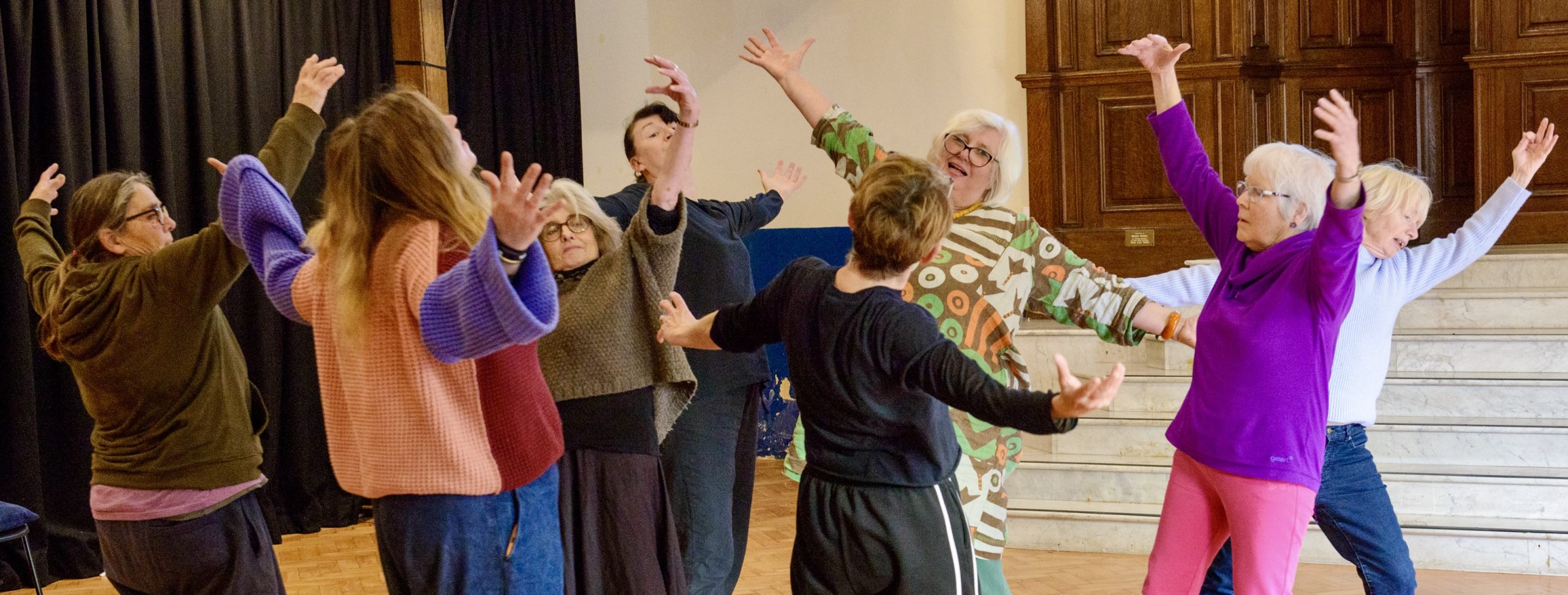 A group of people dressed in colour clothing dance in a hall with arms outstretched.
