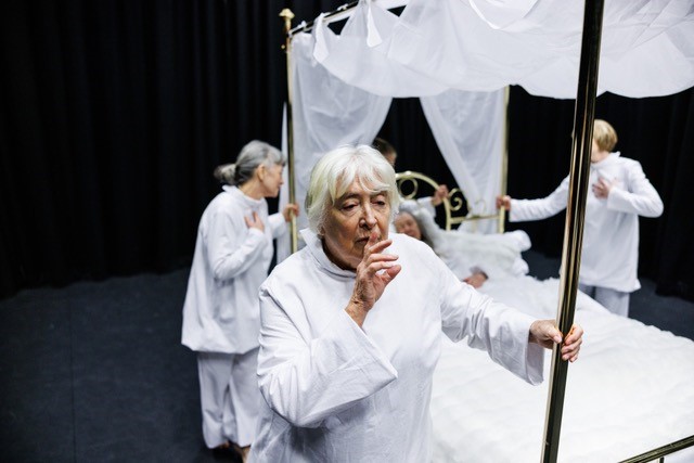 3 women dressed in white push a brass 4 poster bed. A white haired woman in the foreground, holds her finger to her lips.