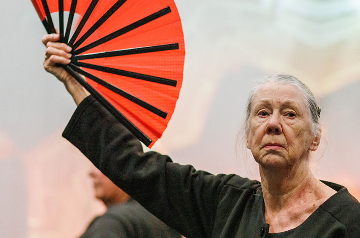 A woman with grey hair tied back, stands staring at the camera and holds a big red fan. She stares at the camera with a grumpy face.