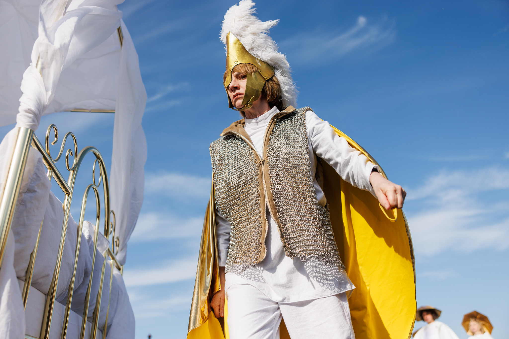 A woman with short brown hair wears a chain mail waistcoat and roman helmet with white feathers. Her fists are clenched and she wears a serious expression. She stands next to a brass four poster bed.