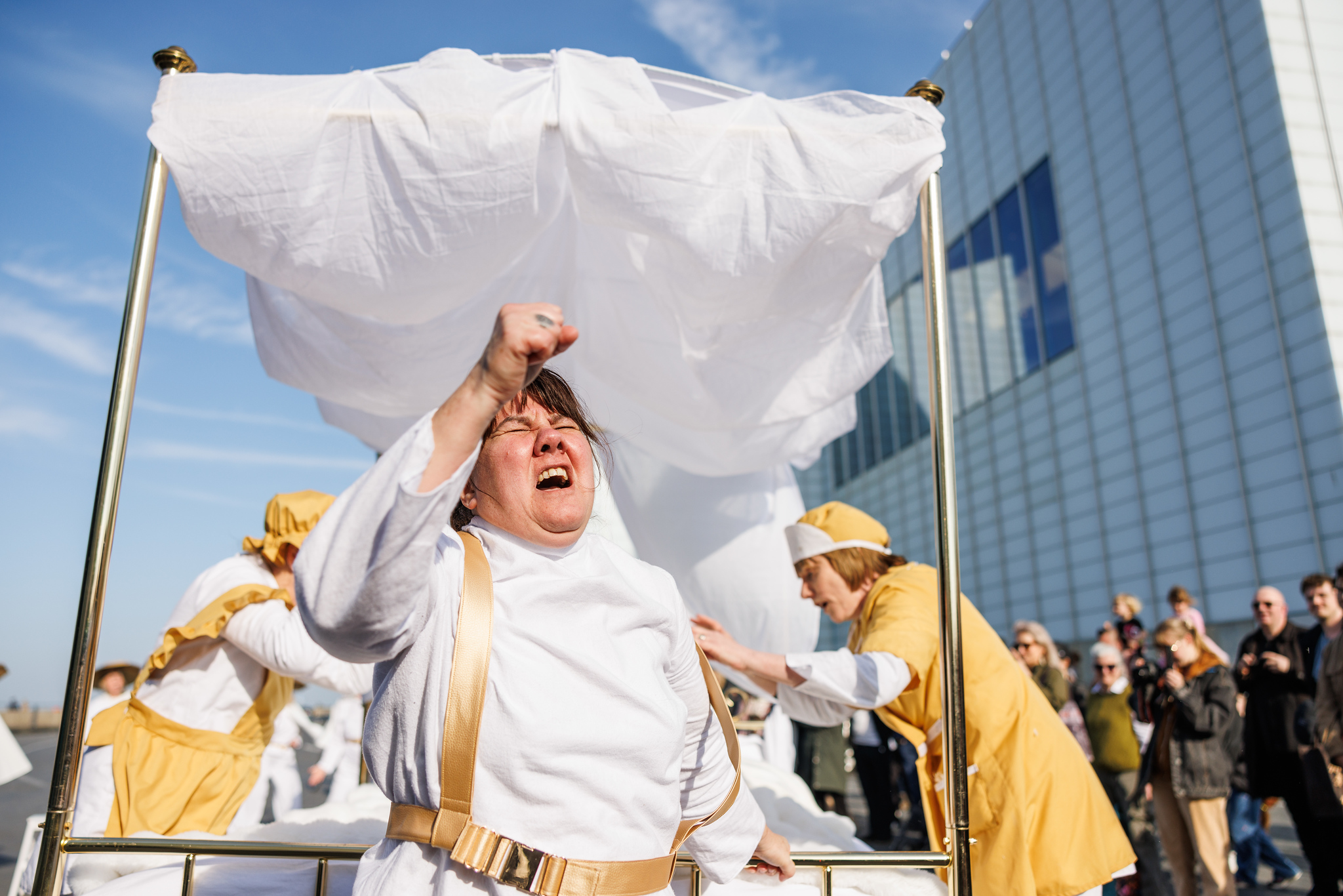 A woman in white with brown hair grimaces with her fist clenched as she pulls a brass four poster bed. Two women dressed as maids are bent over tending to someone in the bed.