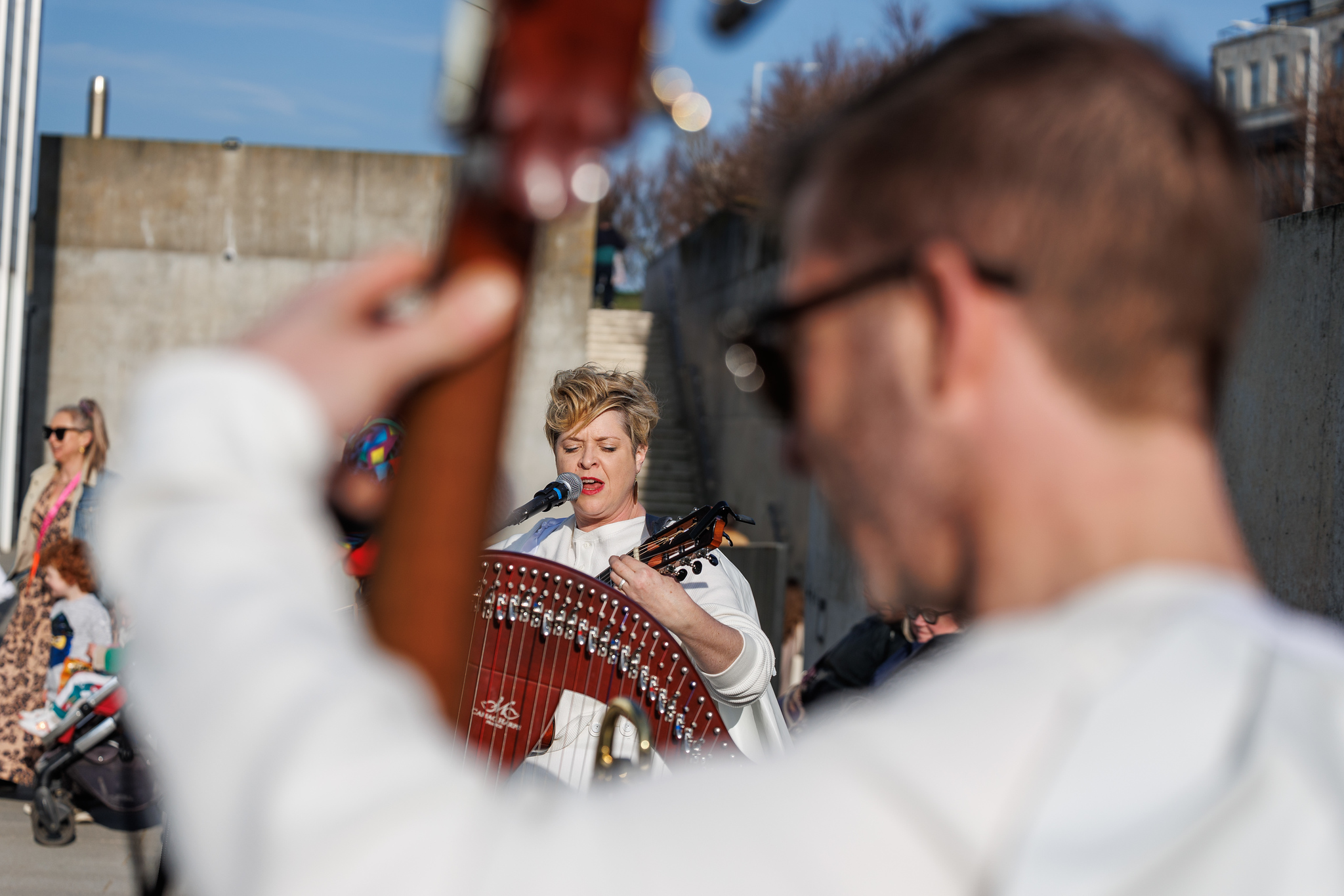 A woman with short blonde hair sings into a microphone while holding a mandolin. In the foreground is the blurred back of a man playing a double bass.