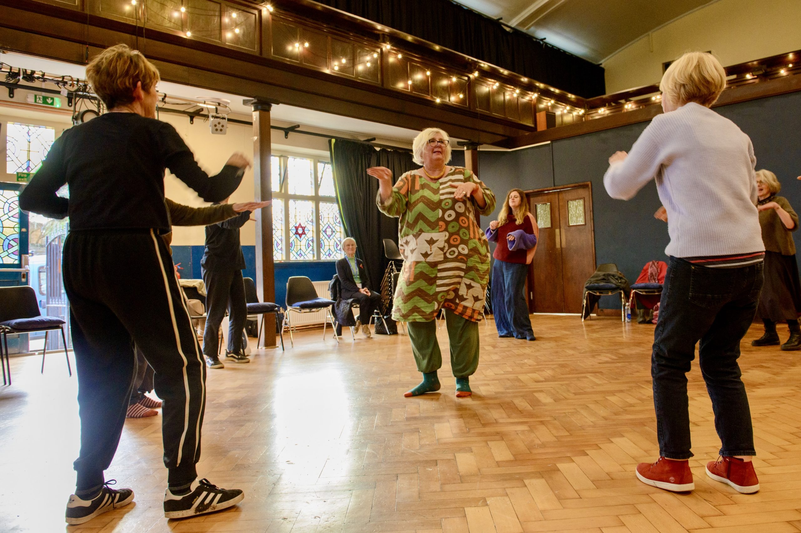 A woman in a multicoloured dress and green trousers with short grey hair stands in the middle of a room dancing, while other people dance around her in a circle.