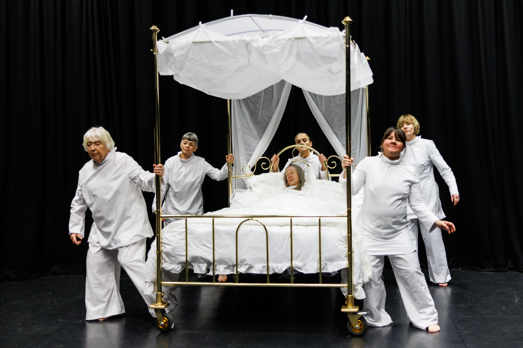 5 women dressed in white, push a brass 4 poster bed while a woman lies in the bed fast asleep under white sheets.