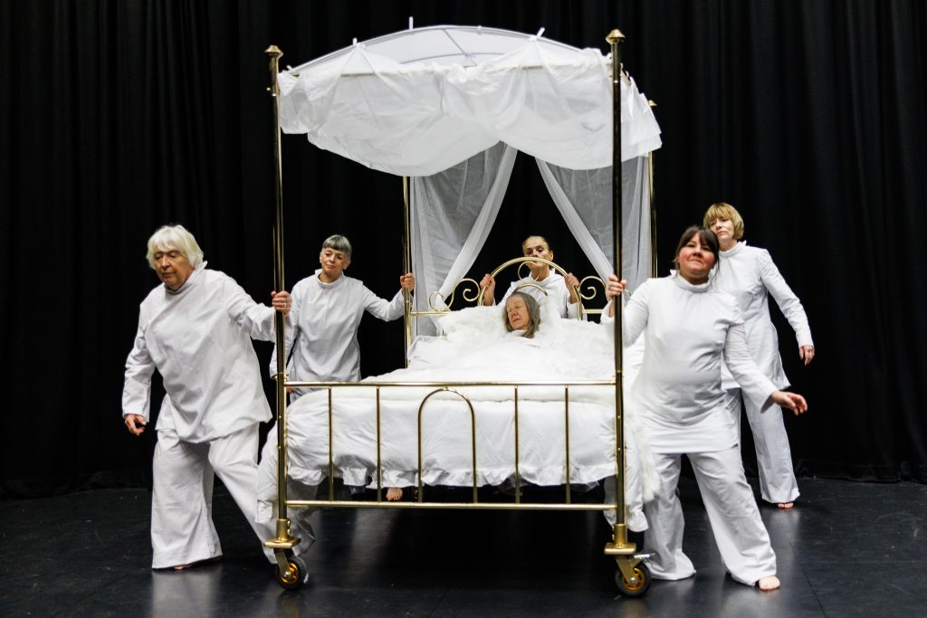 5 women dressed in white, push a brass 4 poster bed while a woman lies in the bed fast asleep under white sheets.