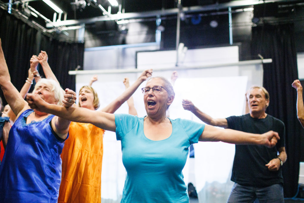A group of older adults in brightly coloured clothing energetically dancing together in a studio, arms raised and smiling in mid-movement.