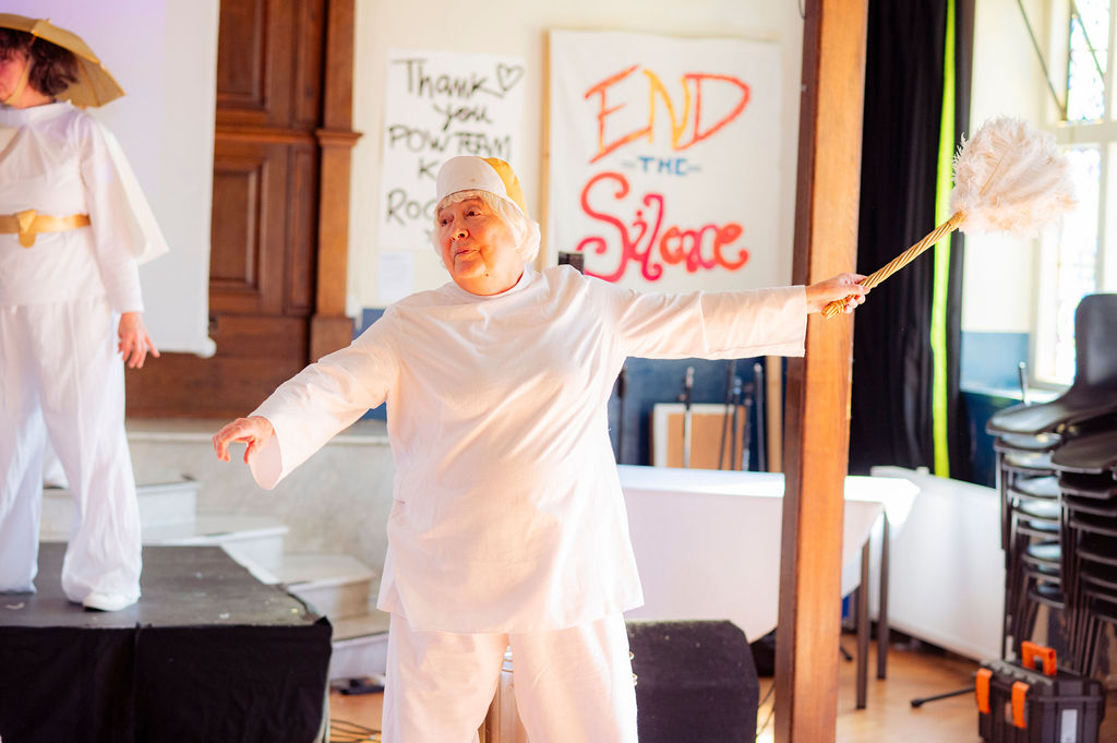 A woman with short white hair stands with an arm outstretched holding a white feather duster. She is wearing a maid's hat and a white top and trousers. There is a sign in the background that says 'End the Silence'.