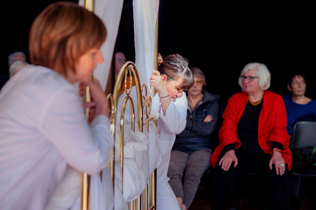 2 women dressed in white push a brass 4 poster bed, while audience members look on.