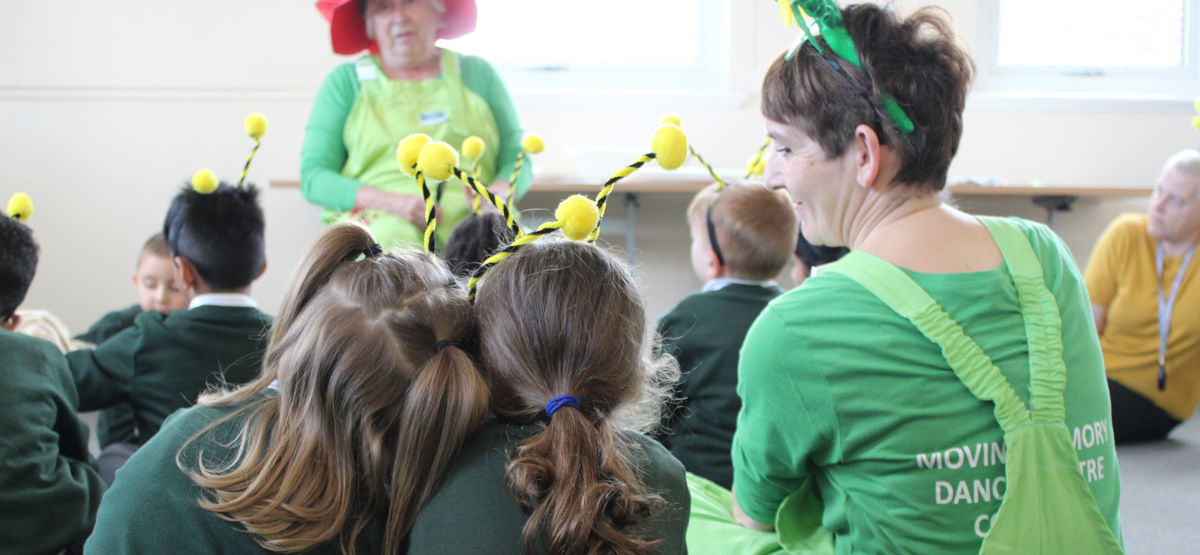 A woman dressed in green dungarees looks and two school children compassionately. They are sitting down with their backs to us.