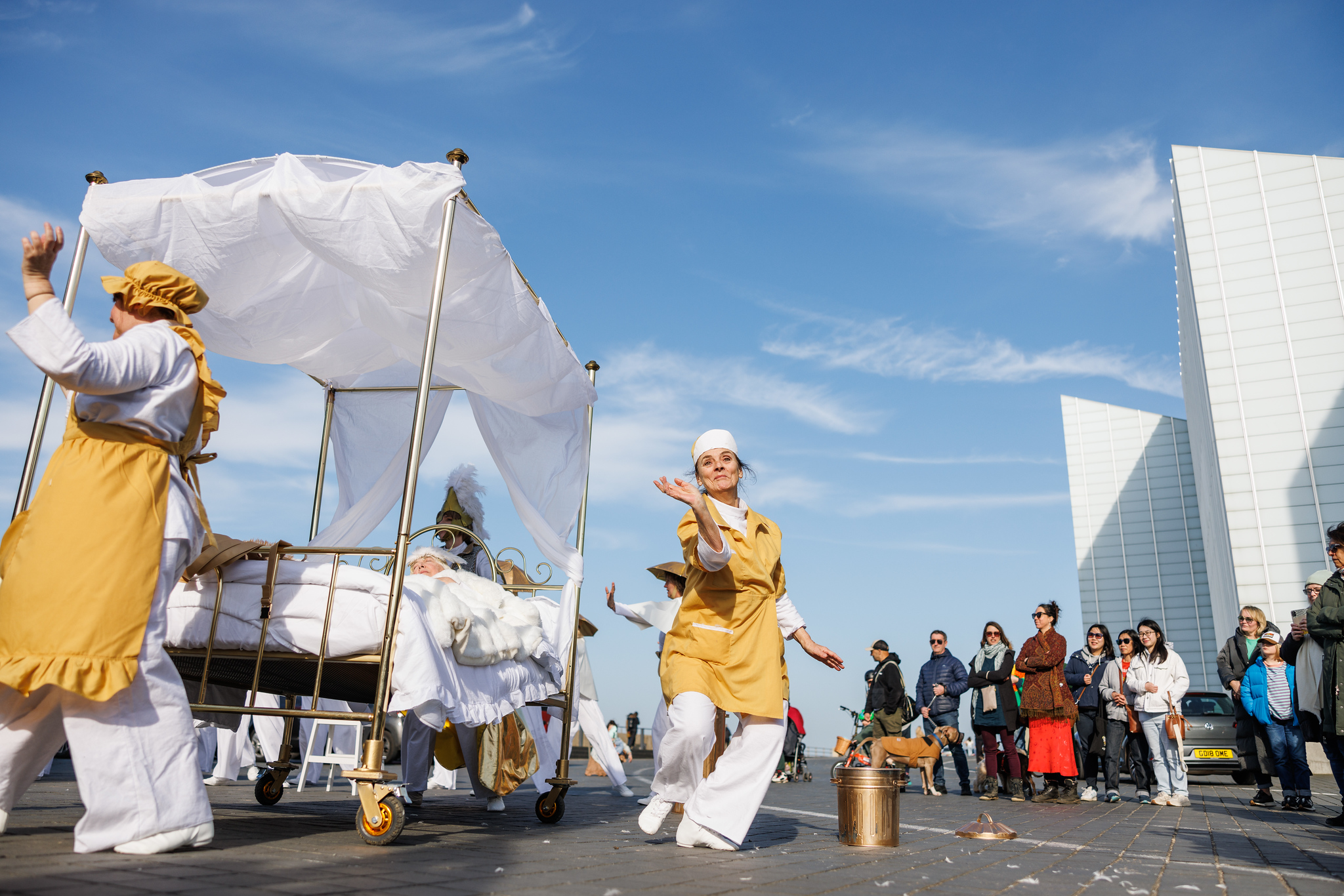 A woman in a yellow maid's outfit dances next to a brass four poster bed.