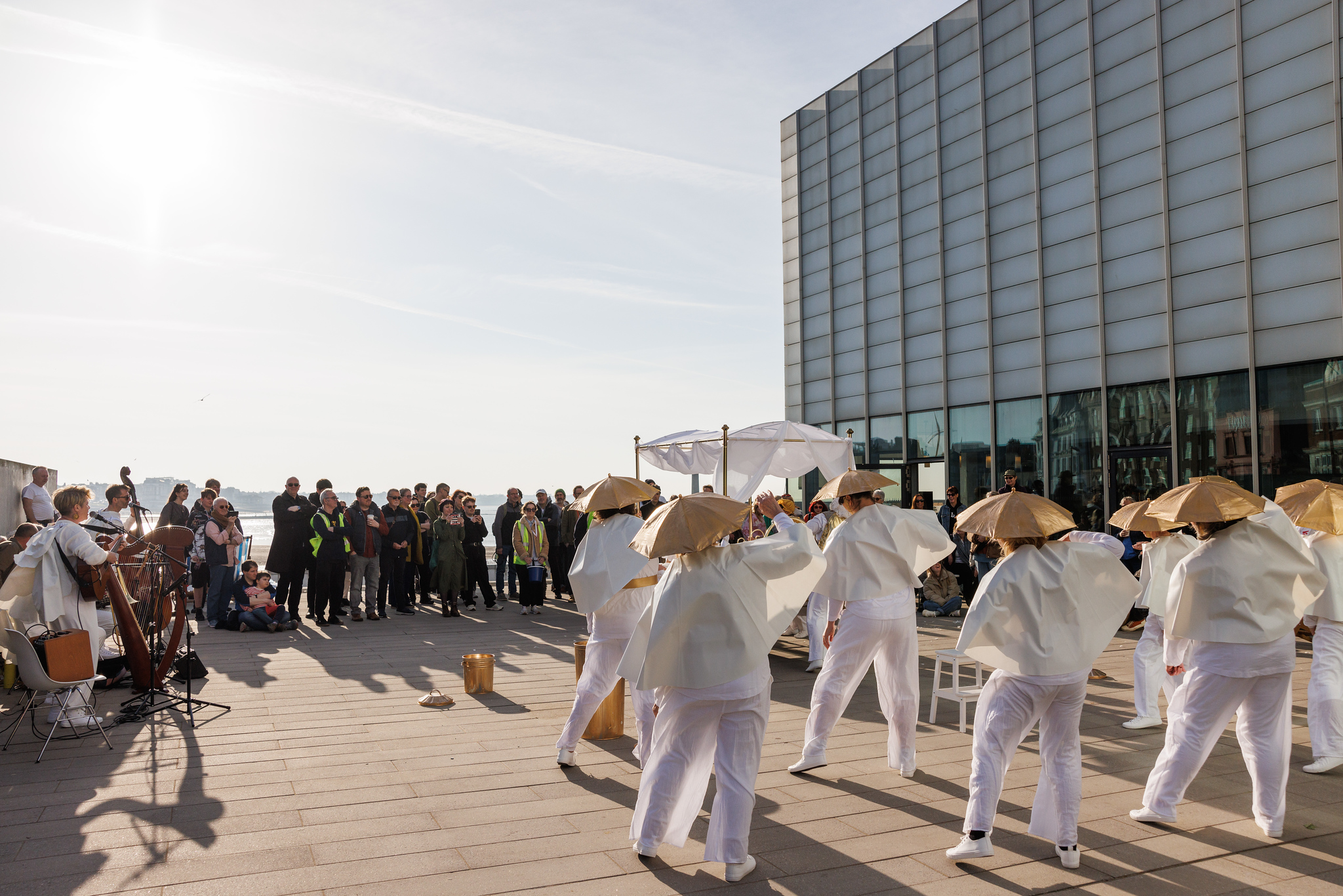 A group of women dressed in white outfits with golden umbrellas on their hands dance with their backs to us but facing an audience who are standing up watching them perform outdoors.