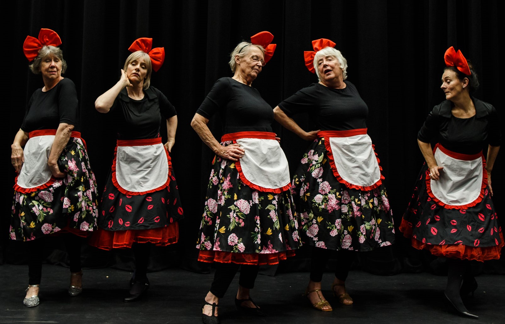 5 women with big red bows in their hair, flowery skirts and white aprons stand in a line and strike a pose.
