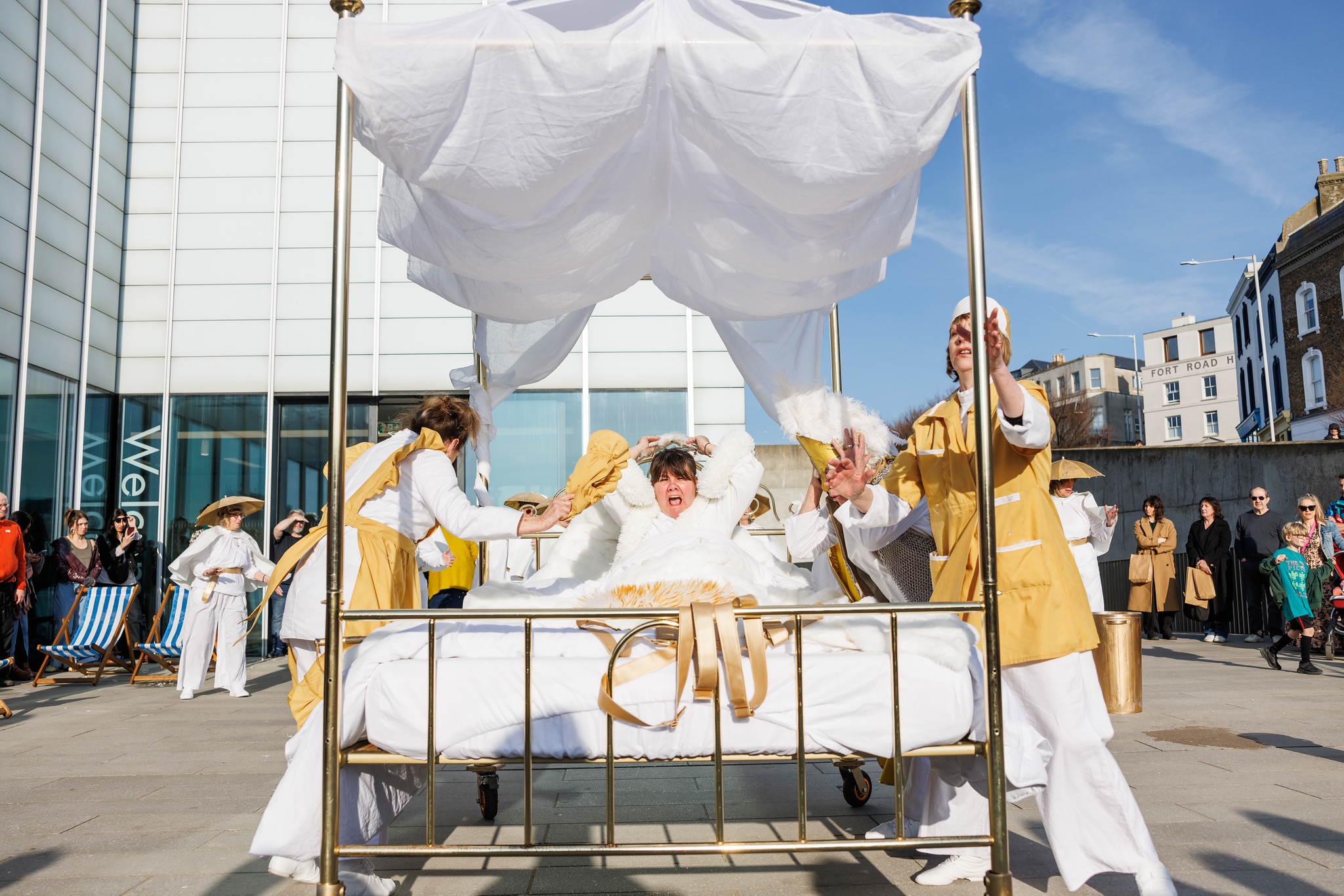 2 women in yellow maids' outfits stand round a brass four poster bed as a woman sits in the bed screaming.