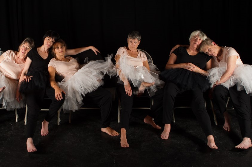 6 women in black outfits and tutus lean on each other and sit on chairs while staring at the camera.