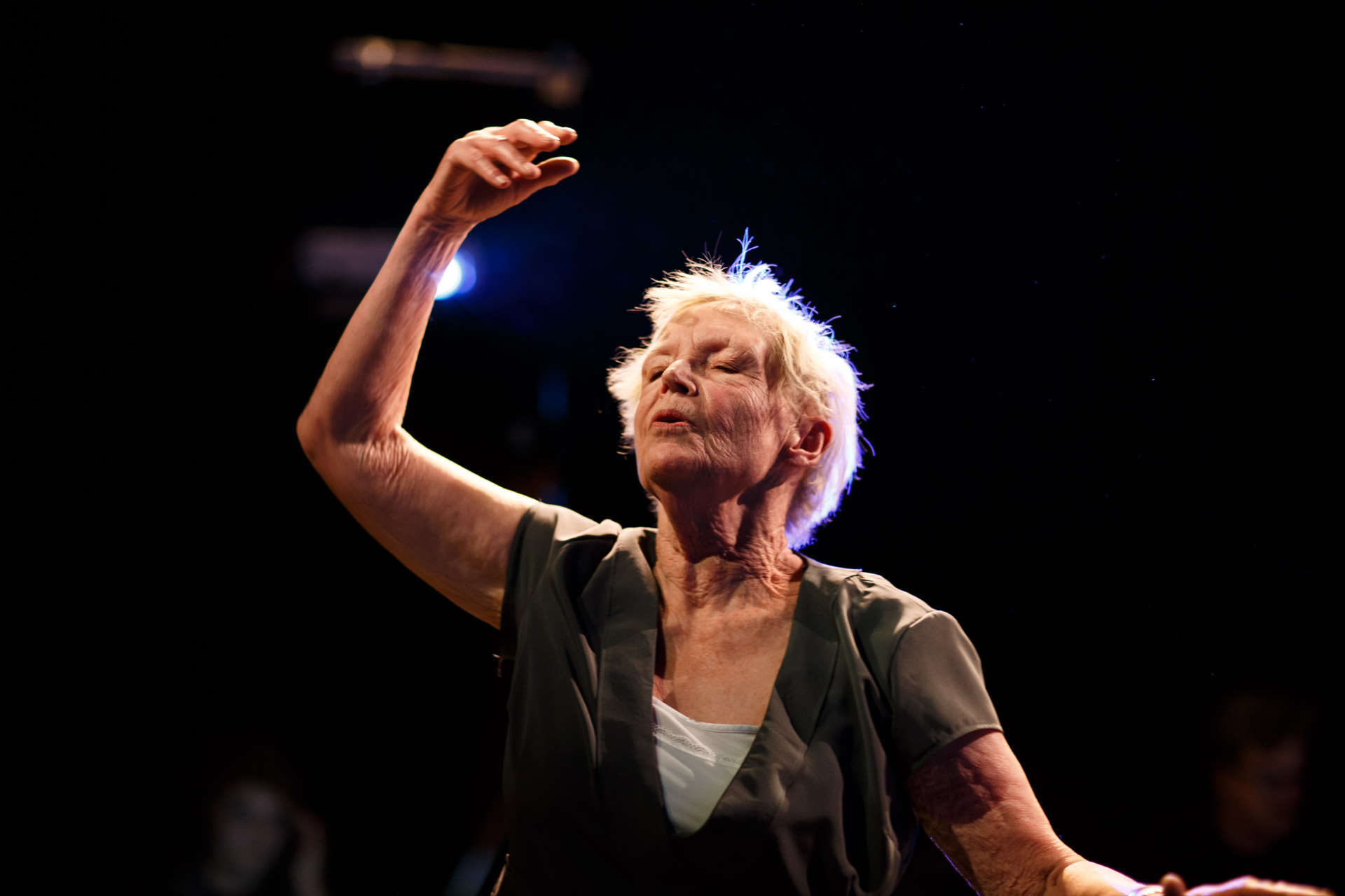 A woman with short white hair and a black top dances with her eyes closed and arm outstretched.