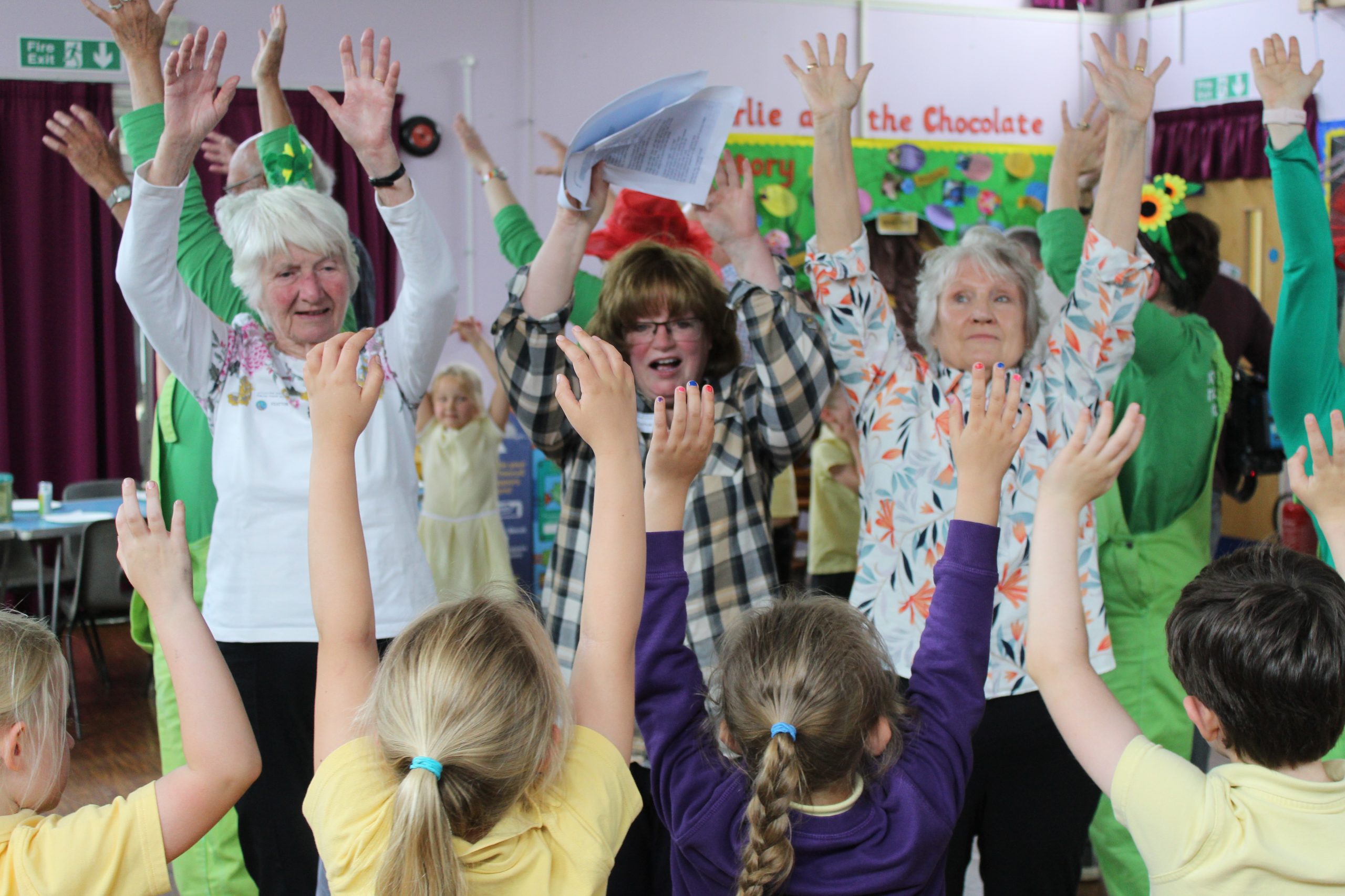 A group of older people stand with their hands in the air. Facing them and with their backs to us are a group of school children, also with their arms in the air.