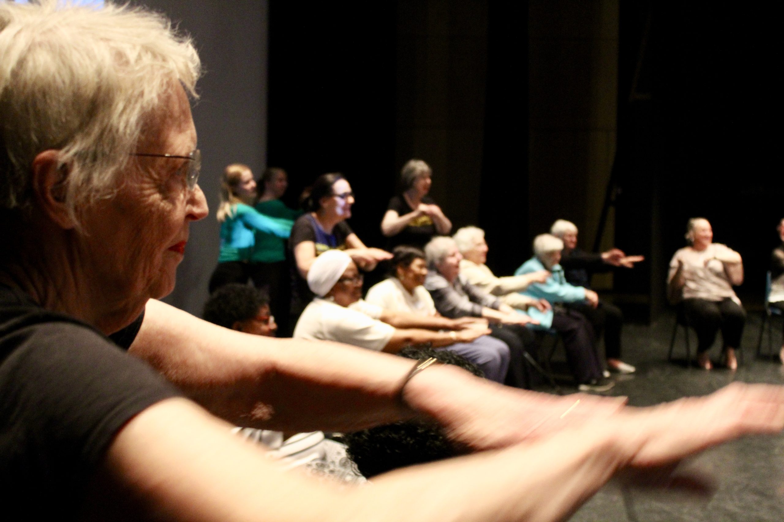 A group of older people sit and stand with their arms outstretched.
