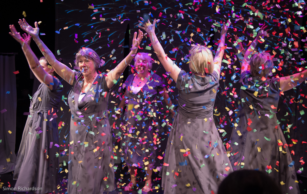 A group of women dressed in black raise their hands in the air in jubilation while confetti falls.