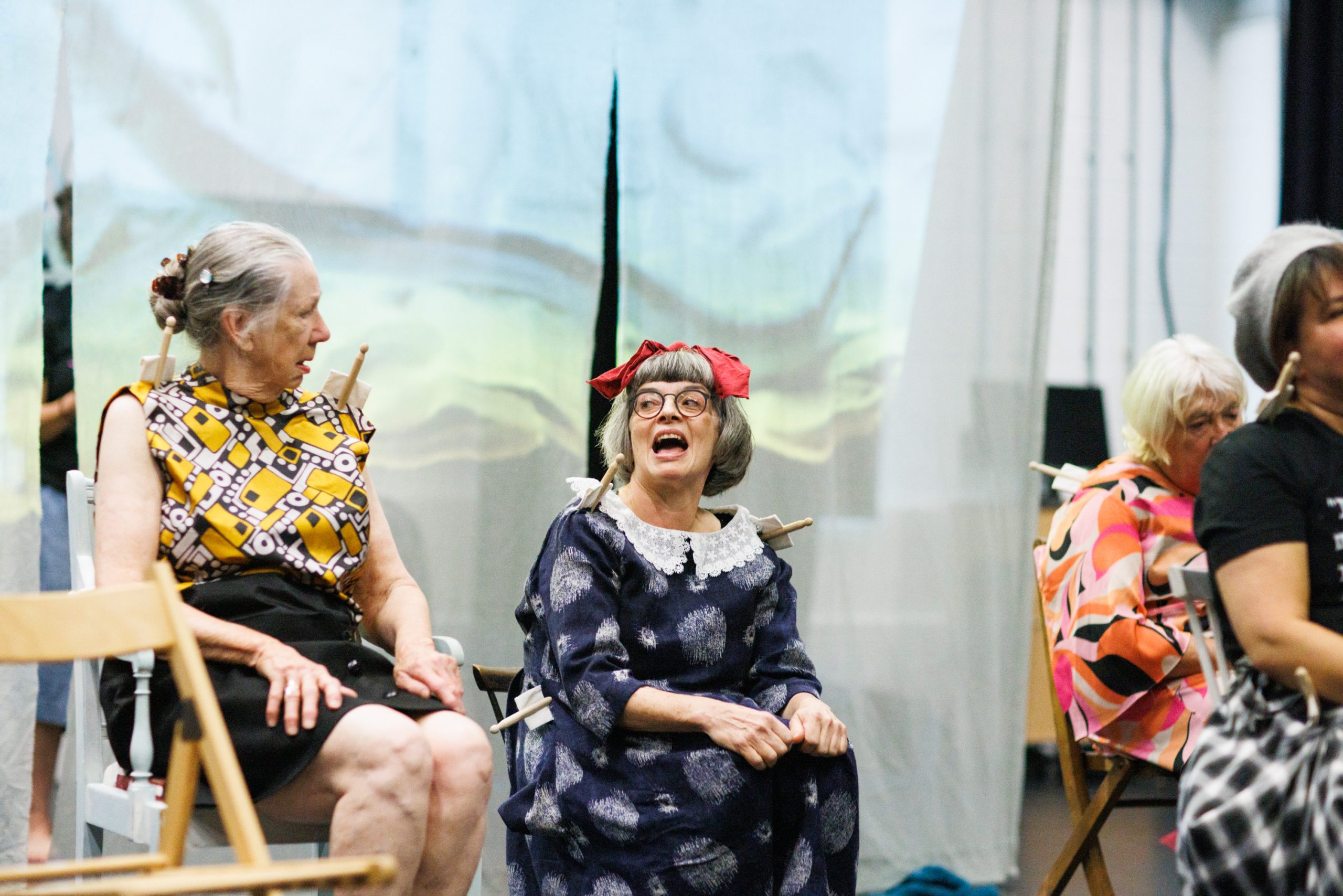 Five women are on a stage. One woman is in the centre, seated, wearing a blue patterned dress, glasses, and a red bow. She is laughing. To her left, another woman with grey hair is seated, wearing a yellow and black patterned top. To the right, a woman in a colourful top is partially visible.