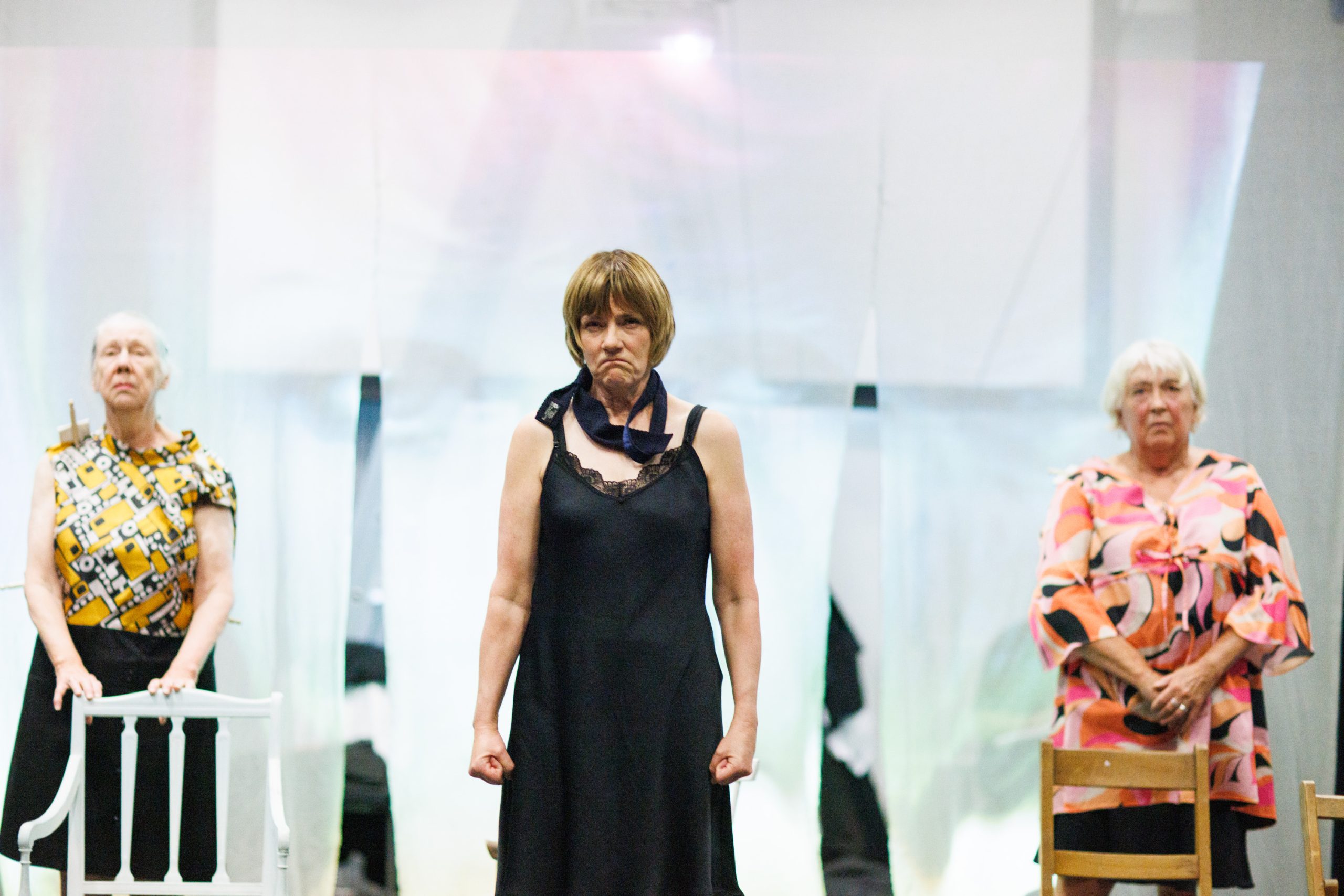 Three women stand on a stage. The woman in the center wears a black night slip and a dark scarf around her neck, with a frustrated expression. The woman on the left has short grey hair and wears a yellow and black patterned top with a black skirt, leaning against a white chair. The woman on the right has short white hair and wears a pink, orange, and white patterned top, standing near a wooden chair.