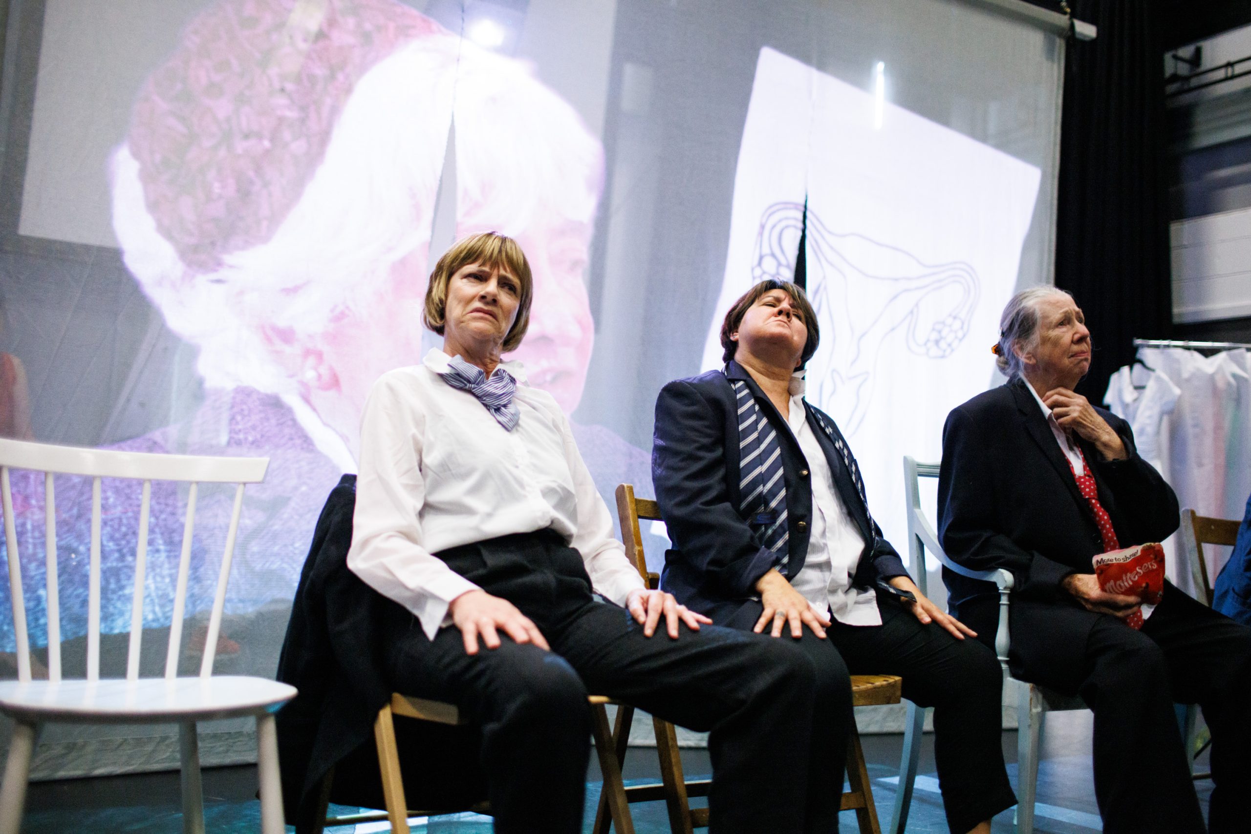 Three women are seated on stage wearing formal men's suits. A blurry image is projected in the background.