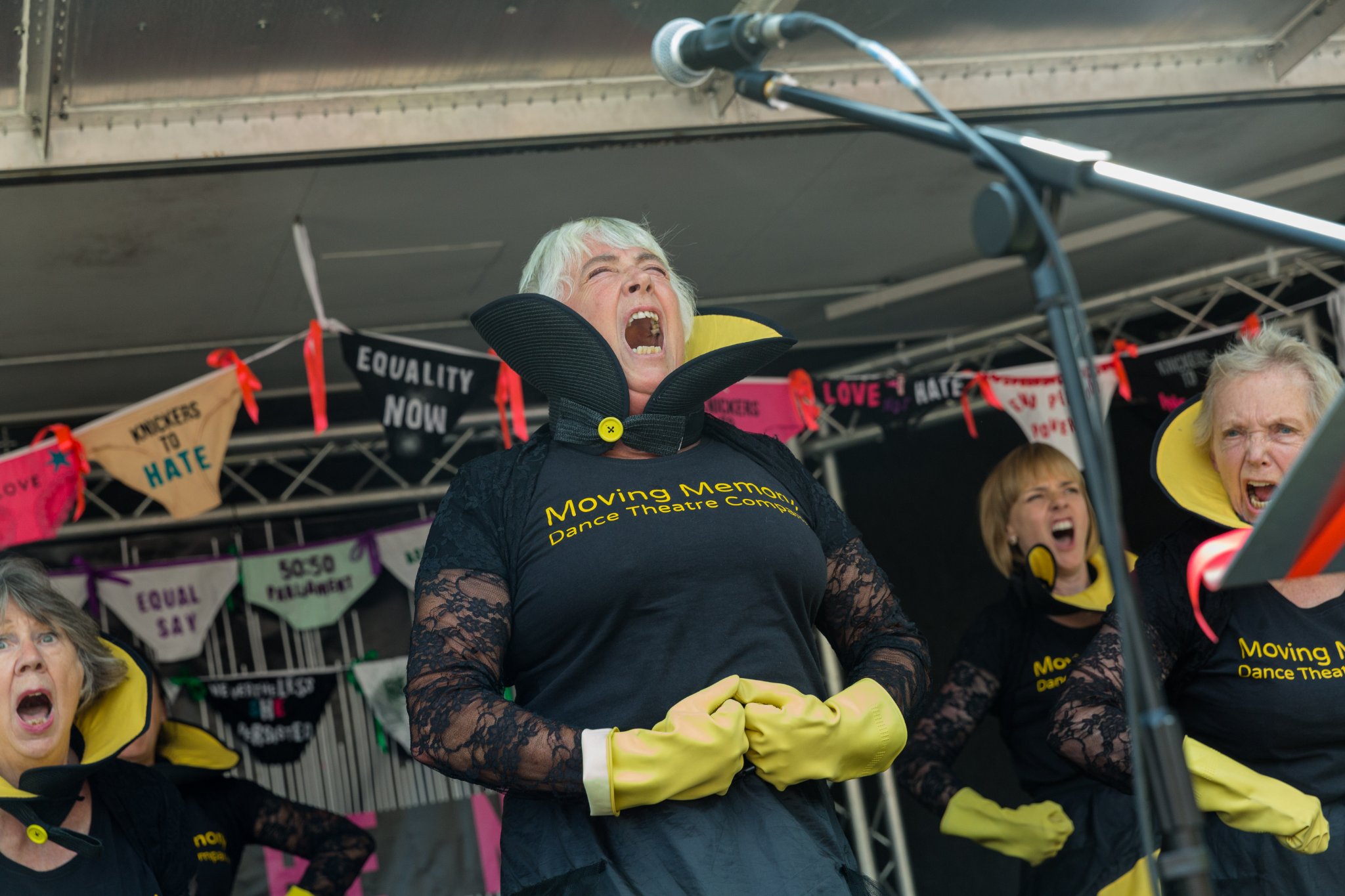 A woman with short grey hair, wearing a black cloak and yellow gloves yells.