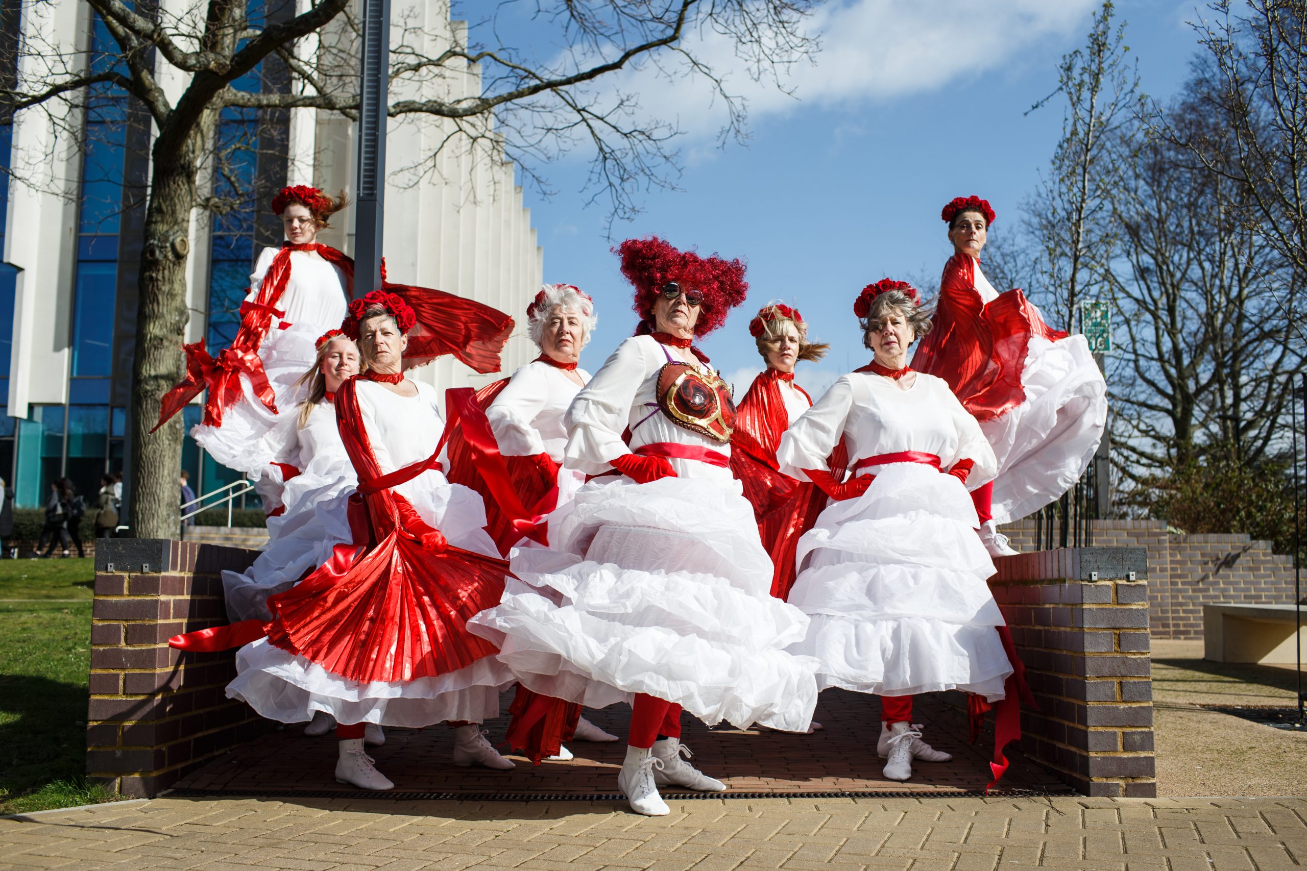 8 women in white dresses with red capes and head dresses stare at the camera with a look of defiance.