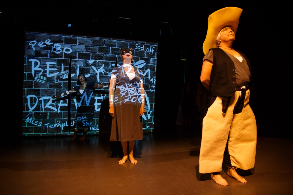 2 women wearing comically large cowboy hats and trousers stand in front of a wall featuring projected words scribbled on.