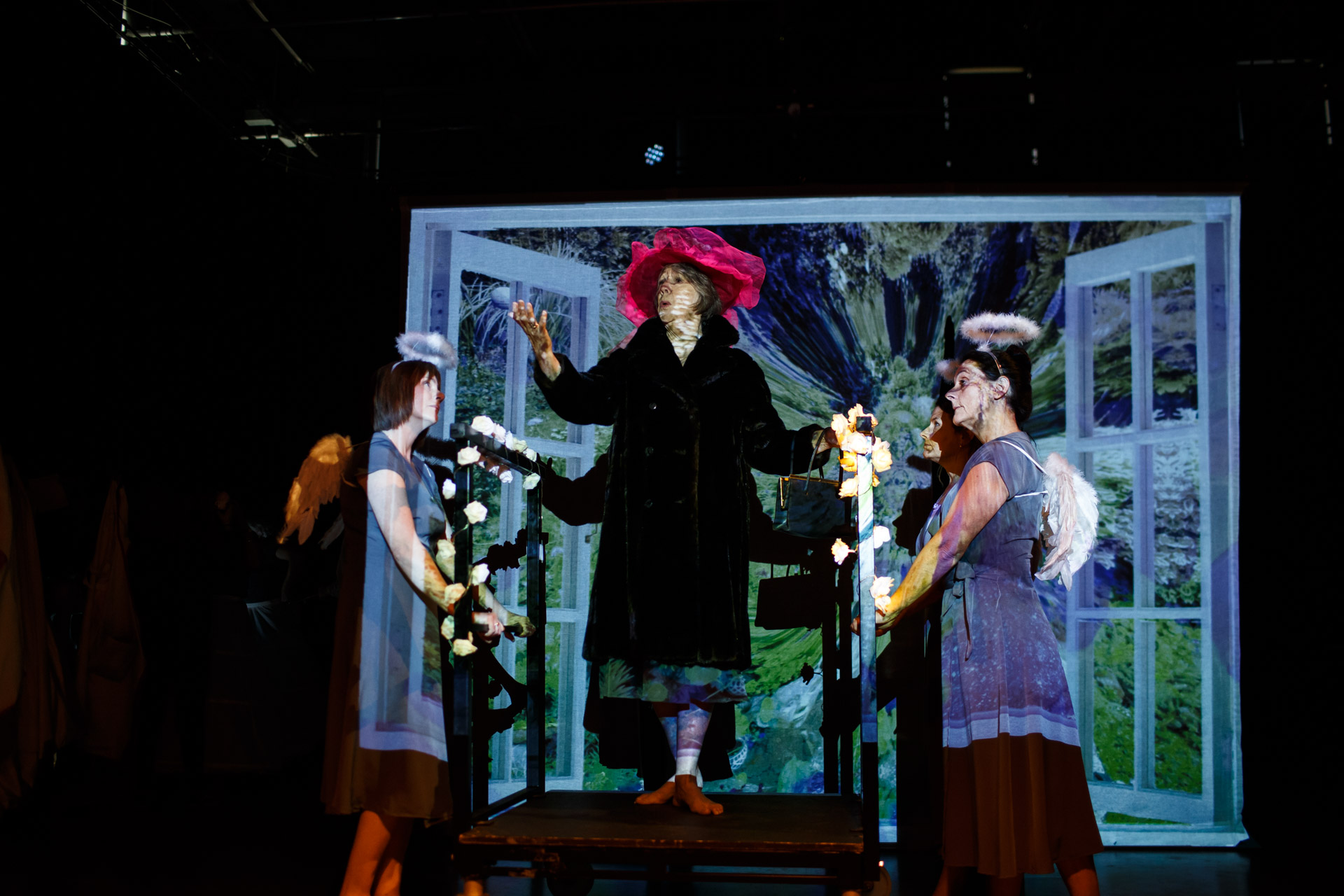 A woman in a red hat addresses the audience as two women in grey dresses wearing angel's halos stand by.