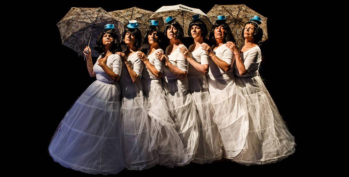 7 women line up in long white cotton dresses, wearing little blue hats and holding parasols.
