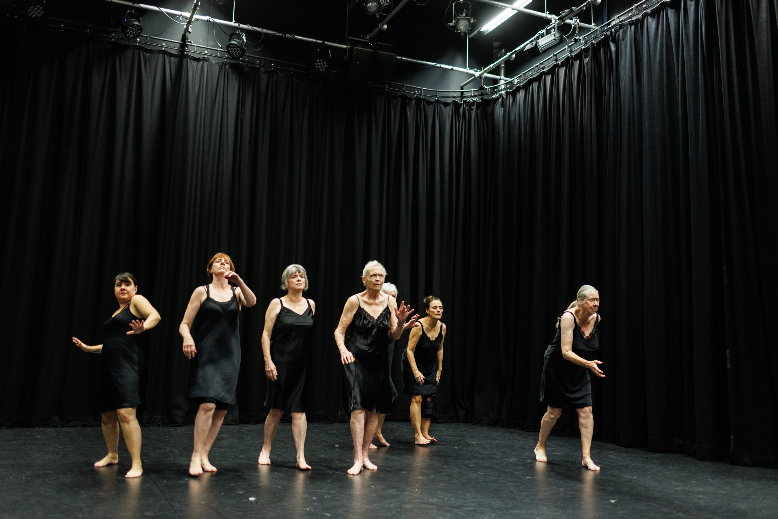 6 women dressed in black night slips walk across a black stage.