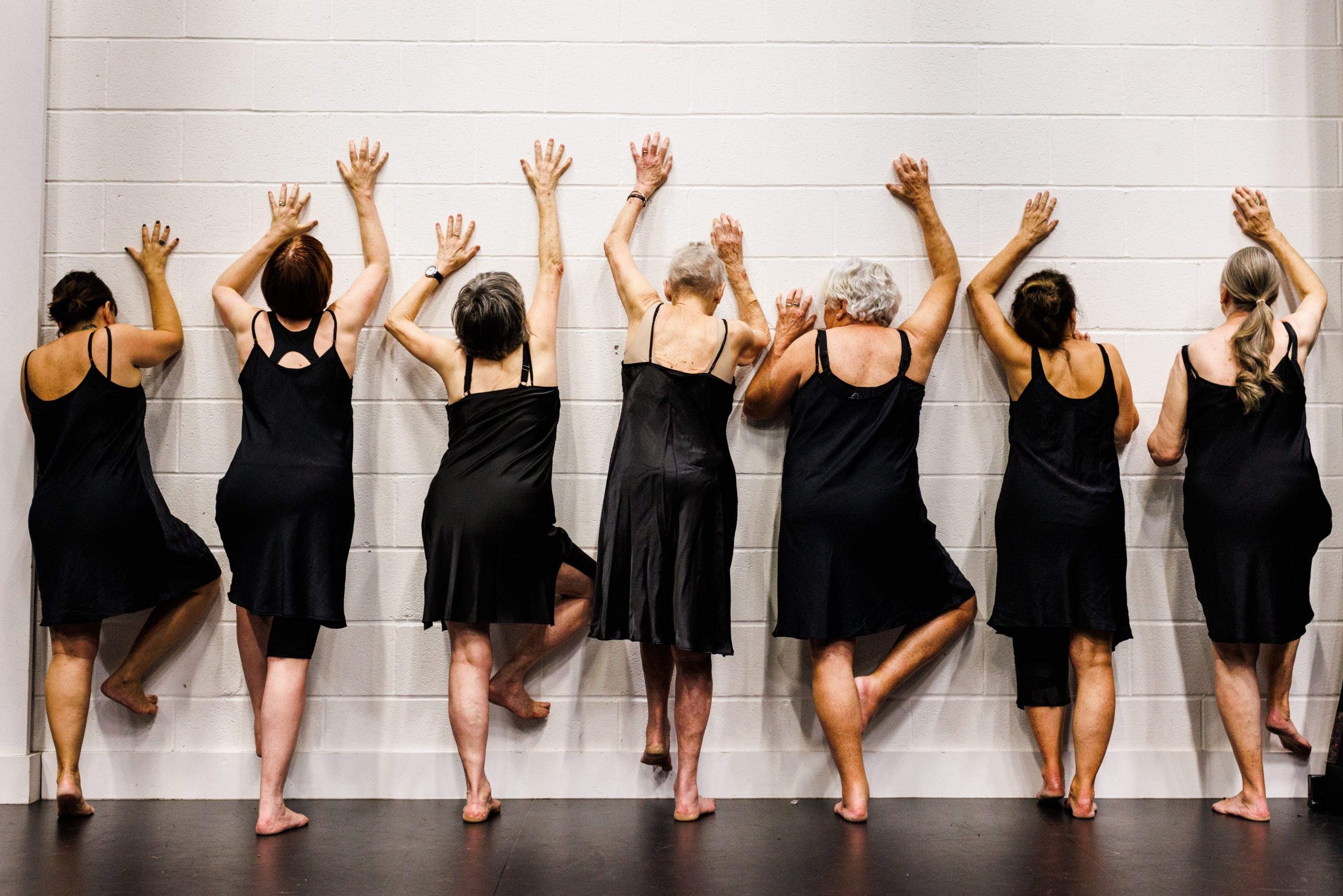 7 women in black slip dresses face a wall with their arms reaching upwards, as if trying to climb up the wall.