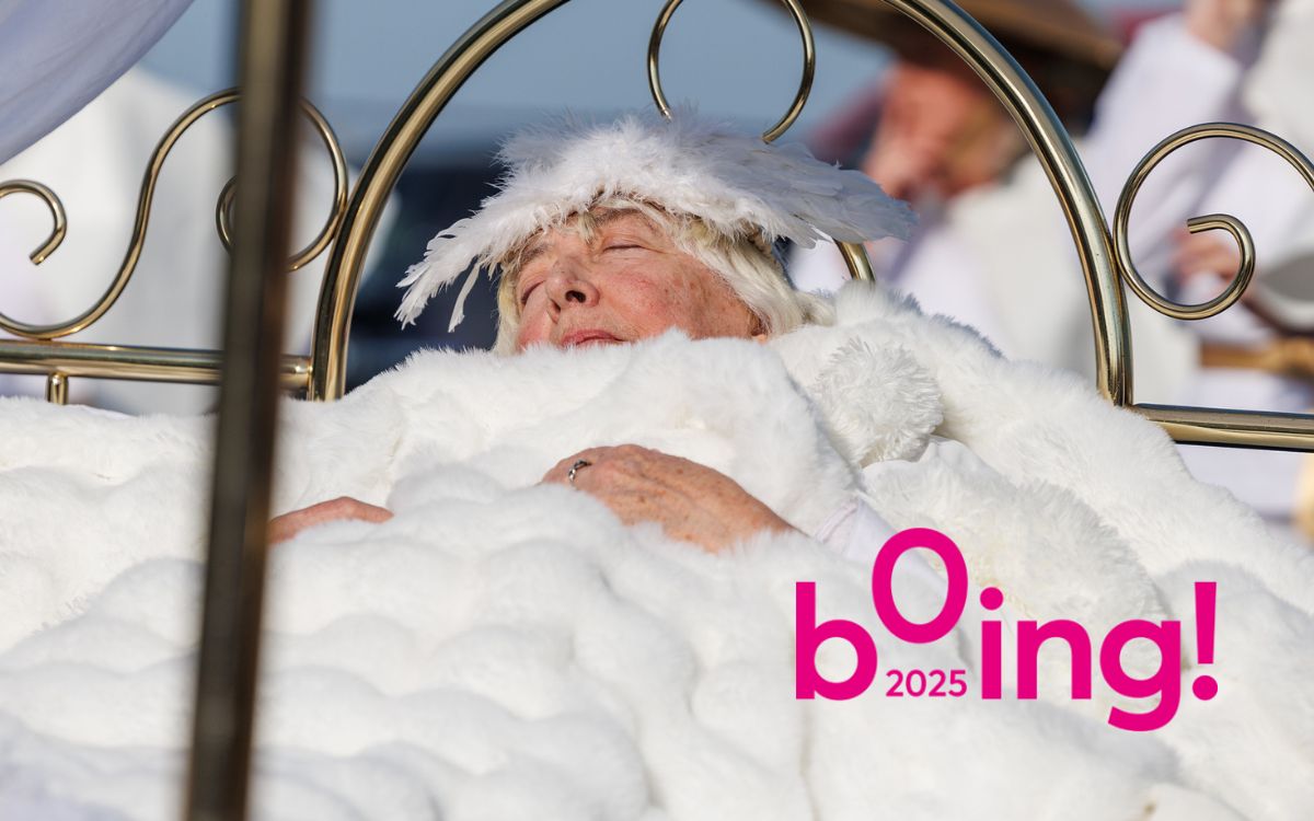 A woman with white hair and a feathered headpiece rests in a golden bed covered with white blankets. A pink BOing! 2025 overlays the image.